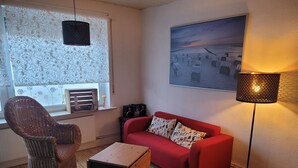 En stue med en rød sofa, en fletstol, et lille træbord, en gulvlampe og et vindue med mønstrede gardiner.