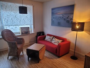 Un salon avec un canapé rouge, un fauteuil en osier, une petite table en bois, un lampadaire et une fenêtre ornée de rideaux à motifs.