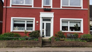 Une maison à deux étages, aux murs de briques rouges, avec des fenêtres à cadre blanc, une porte d’entrée et un petit jardin agrémenté de plantes et d’un mur en briques.