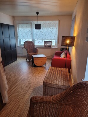 A room with a wooden floor, a red sofa, a wicker chair, a small table, a black lamp, and a window with patterned curtains.