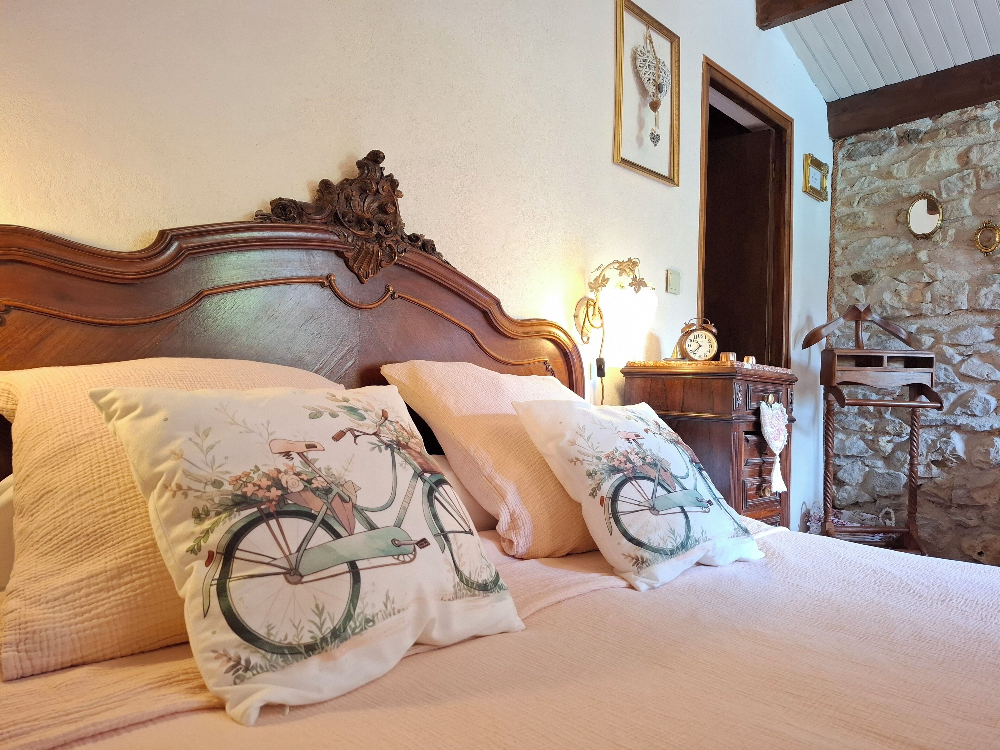 Un dormitorio con cabecera de madera, cama con almohadas estampadas, mesita de noche con un reloj y pared de piedra.