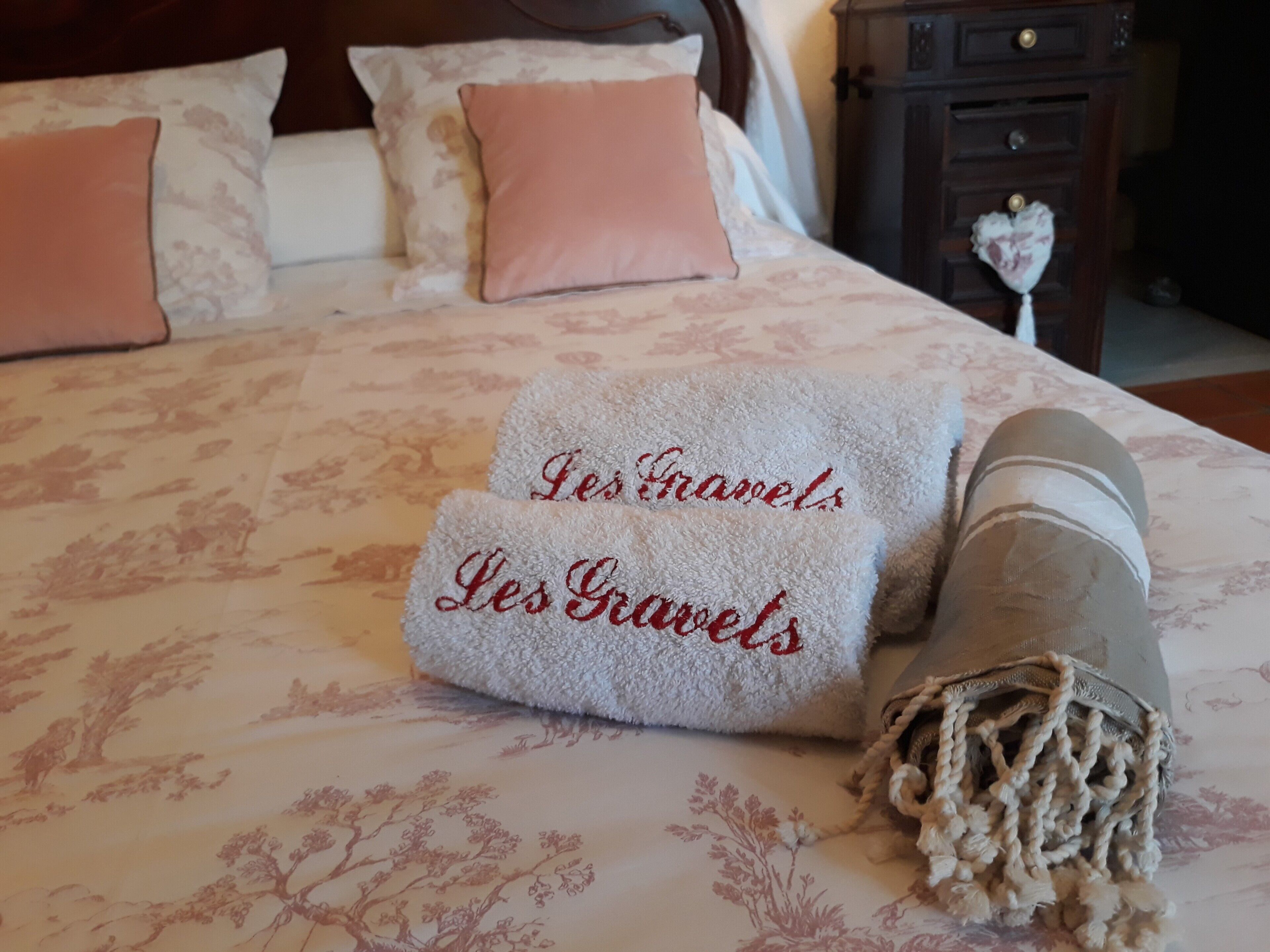 Un lit bien fait, avec une literie à motifs floraux, deux serviettes brodées avec le mot « Les Grands », et une serviette enroulée, maintenue par une cordelette.