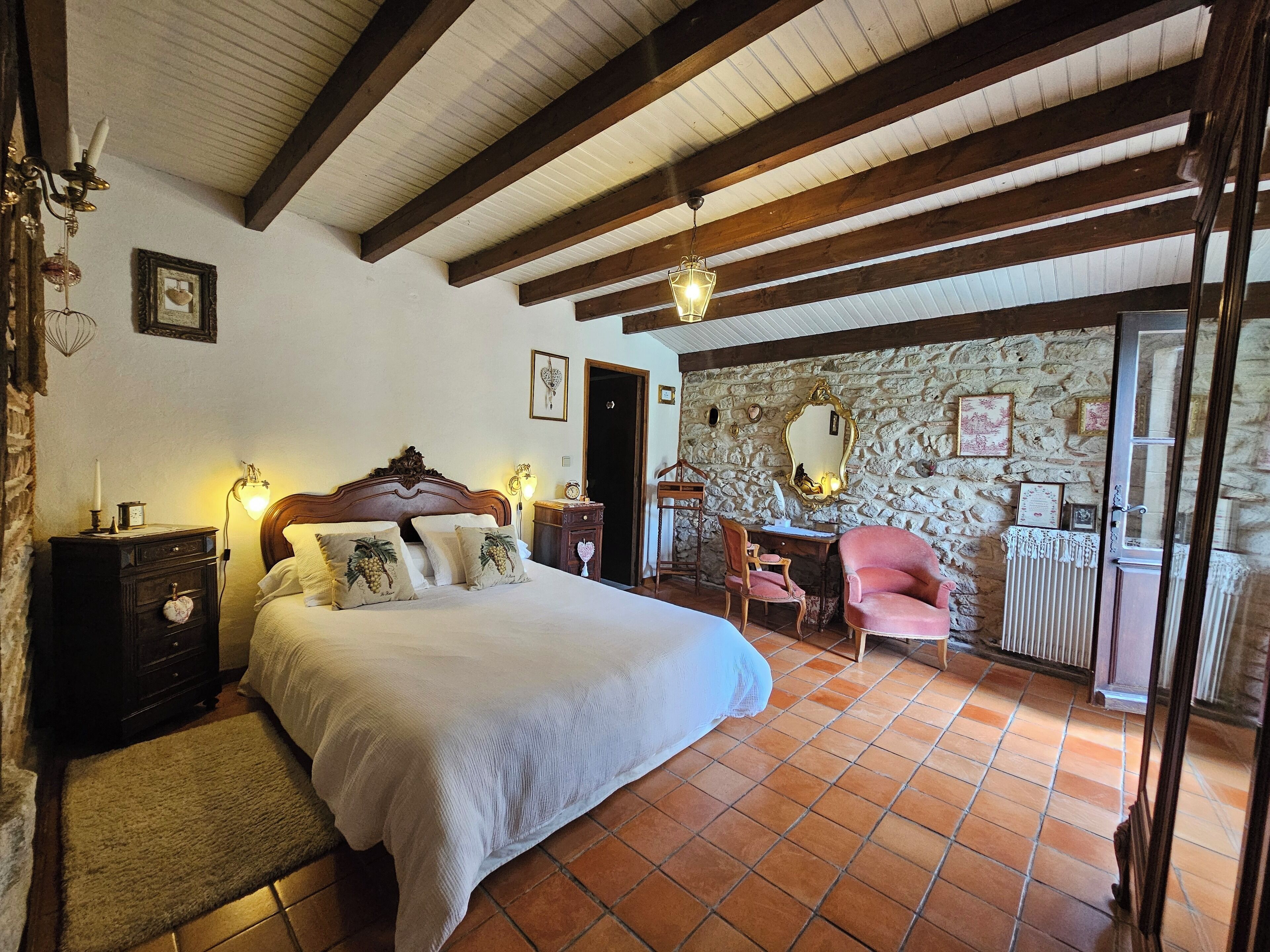 Une chambre avec un grand lit, des poutres en bois, un mur en pierre et un bureau avec une chaise.