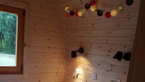 Acogedora habitación tipo cabaña con interior de madera, una cama con ropa blanca, un ventanal y luces colgantes en el techo.