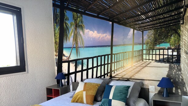 A bedroom with a bed, pillows, a nightstand, and a view of a beach and ocean through a window. - BARCARESA-LA PERLE SEA VIEW WATERFRONT AIR CONDITIONING WIFI (Port Leucate)