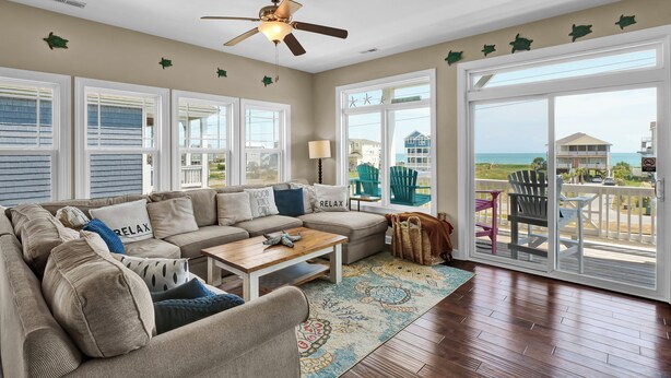 A spacious living room with a large sectional sofa, a ceiling fan, and a view of the ocean through large sliding glass doors. - Linens included - Amazing Views, Steps to Beach, Elevator and Fenced Yard (North Topsail Beach)