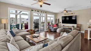 A spacious living room with a large sectional sofa, a wooden coffee table, and a flat-screen TV mounted on the wall. There are two ceiling fans, a large window with a view of the neighborhood, and a sliding glass door leading to an outdoor patio. - Linens included - Amazing Views, Steps to Beach, Elevator and Fenced Yard (North Topsail Beach)