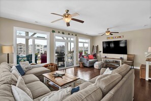 A spacious living room with a large sectional sofa, a wooden coffee table, and a flat-screen TV mounted on the wall. There are two ceiling fans, a large window with a view of the neighborhood, and a sliding glass door leading to an outdoor patio. - Linens included - Amazing Views, Steps to Beach, Elevator and Fenced Yard (North Topsail Beach)
