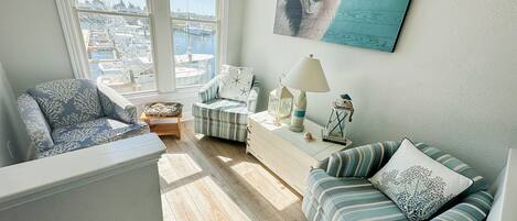 En hyggelig stue med udsigt over en lystbådehavn, med en stribet sofa, en lænestol og et hvidt sofabord med dekorative genstande.