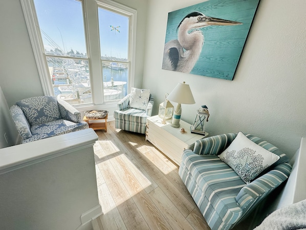 A cozy living room with a view of a marina, featuring a striped sofa, a chair, and a white side table with decorative items.
