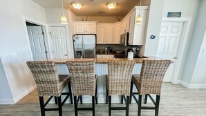 A kitchen with white cabinets, a stainless steel refrigerator, and a dining area with wicker chairs and a central island. - Offshore Addiction  (Manteo)