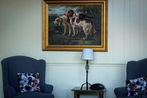 A room with two armchairs, each with a dog-themed pillow, a small table with a lamp, and a framed painting of two dogs on the wall.