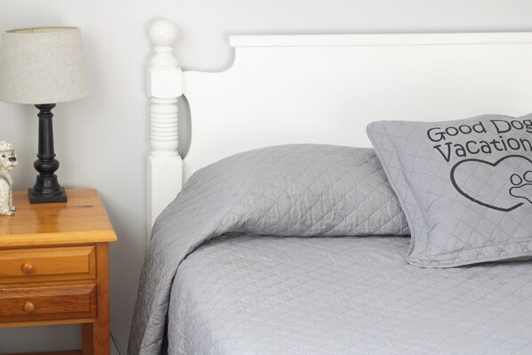 A bed with a quilted grey blanket, a pillow with 'Good Dog Vacation' text, a white headboard, a wooden nightstand with a lamp, and a small dog figurine.
