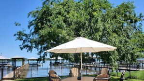 An outdoor patio with a table and chairs under a canopy, overlooking a body of water with docks and a fence.