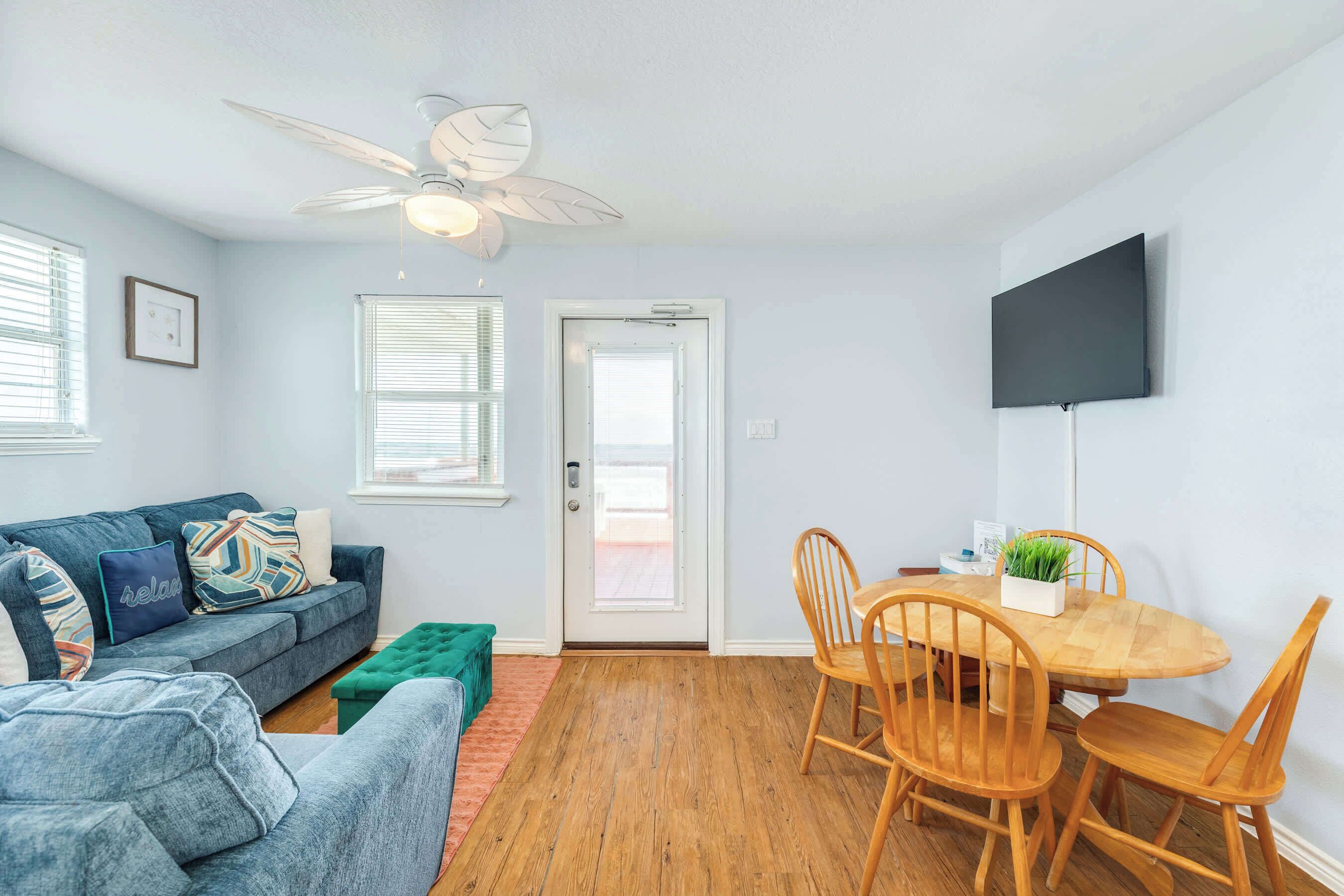 A living room with a sofa, a dining table with chairs, a ceiling fan, and a TV mounted on the wall.