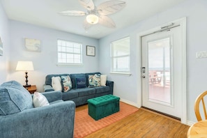 A living room with a sofa, ottoman, ceiling fan, and two windows with blinds.