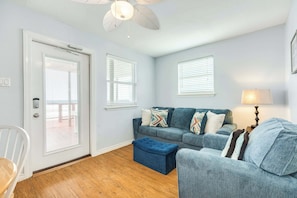 A living room with a blue sofa, a matching ottoman, a ceiling fan, and a door leading to an outdoor area.
