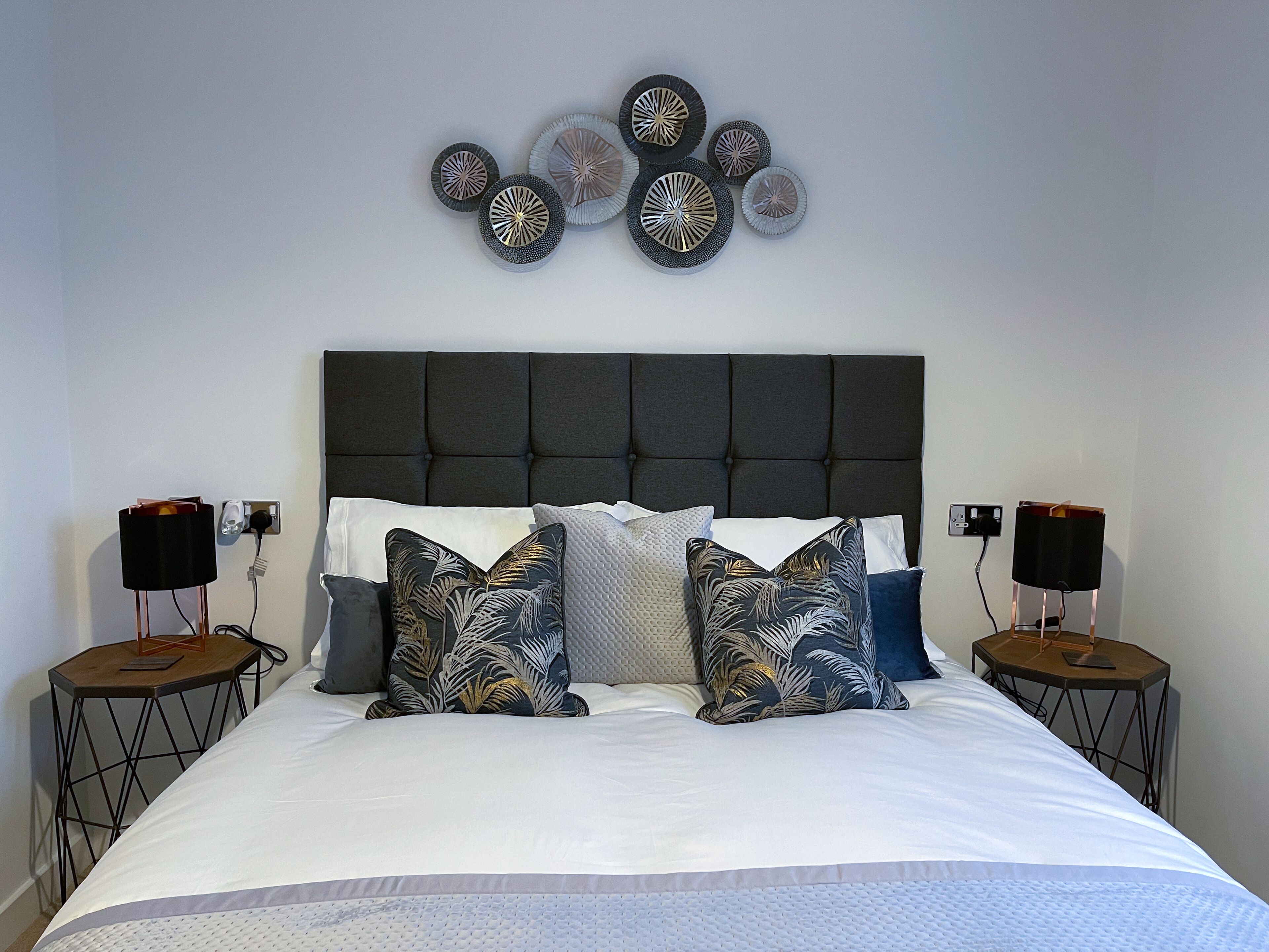 A neatly made bed with a tufted headboard, flanked by two nightstands with speakers, and a wall art piece above.