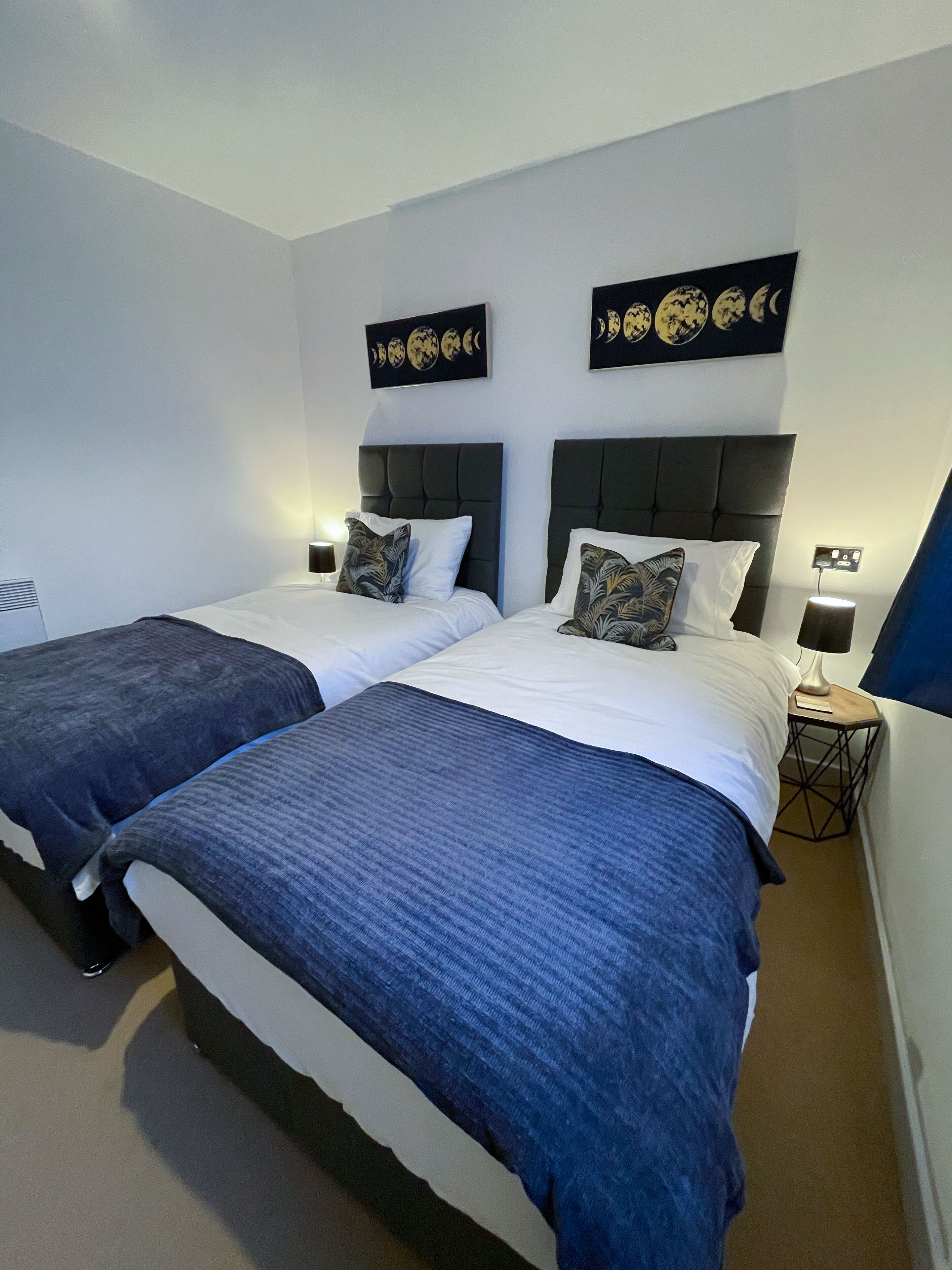Ein Hotelzimmer mit zwei Betten, jedes mit einer dunkelblauen Tagesdecke und weißen Kissen. An der Wand über den Betten befinden sich Kunstwerke, die die Mondphasen darstellen.