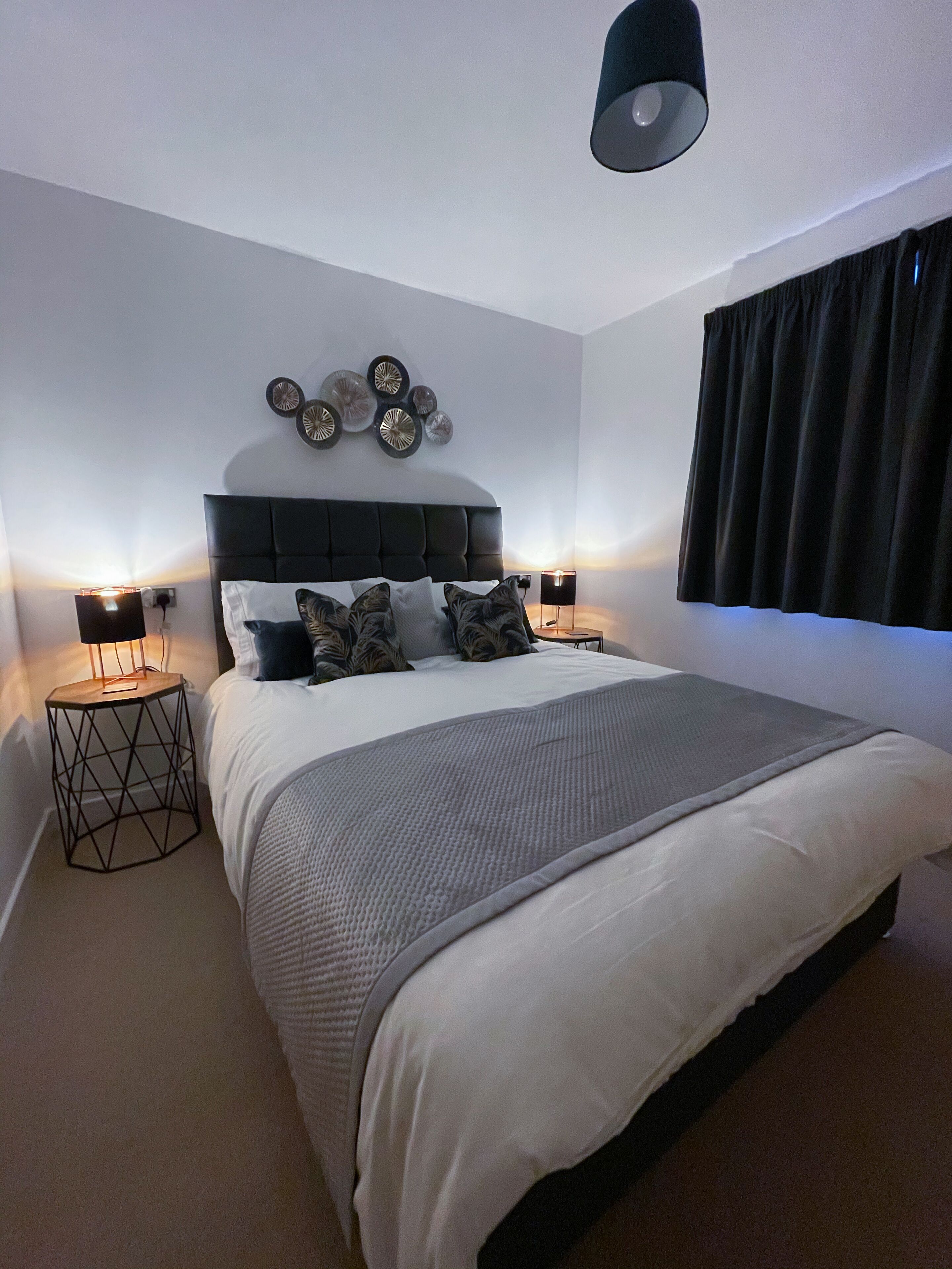 A modern bedroom with a large bed, two bedside tables with lamps, a headboard with decorative plates, and a window with curtains.