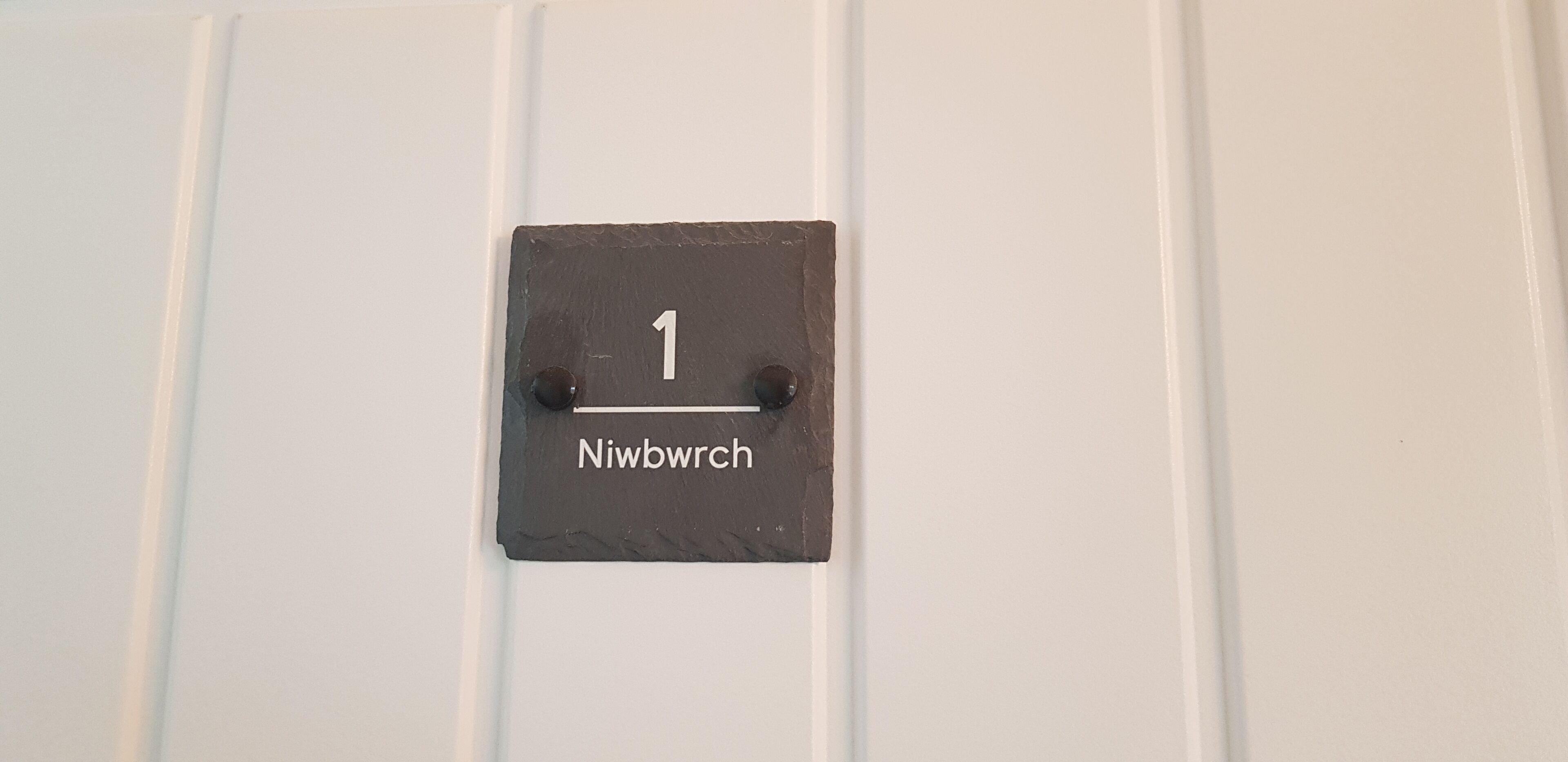 A black slate sign with the number 1 and the word "Niwbwrch" on a white wall.