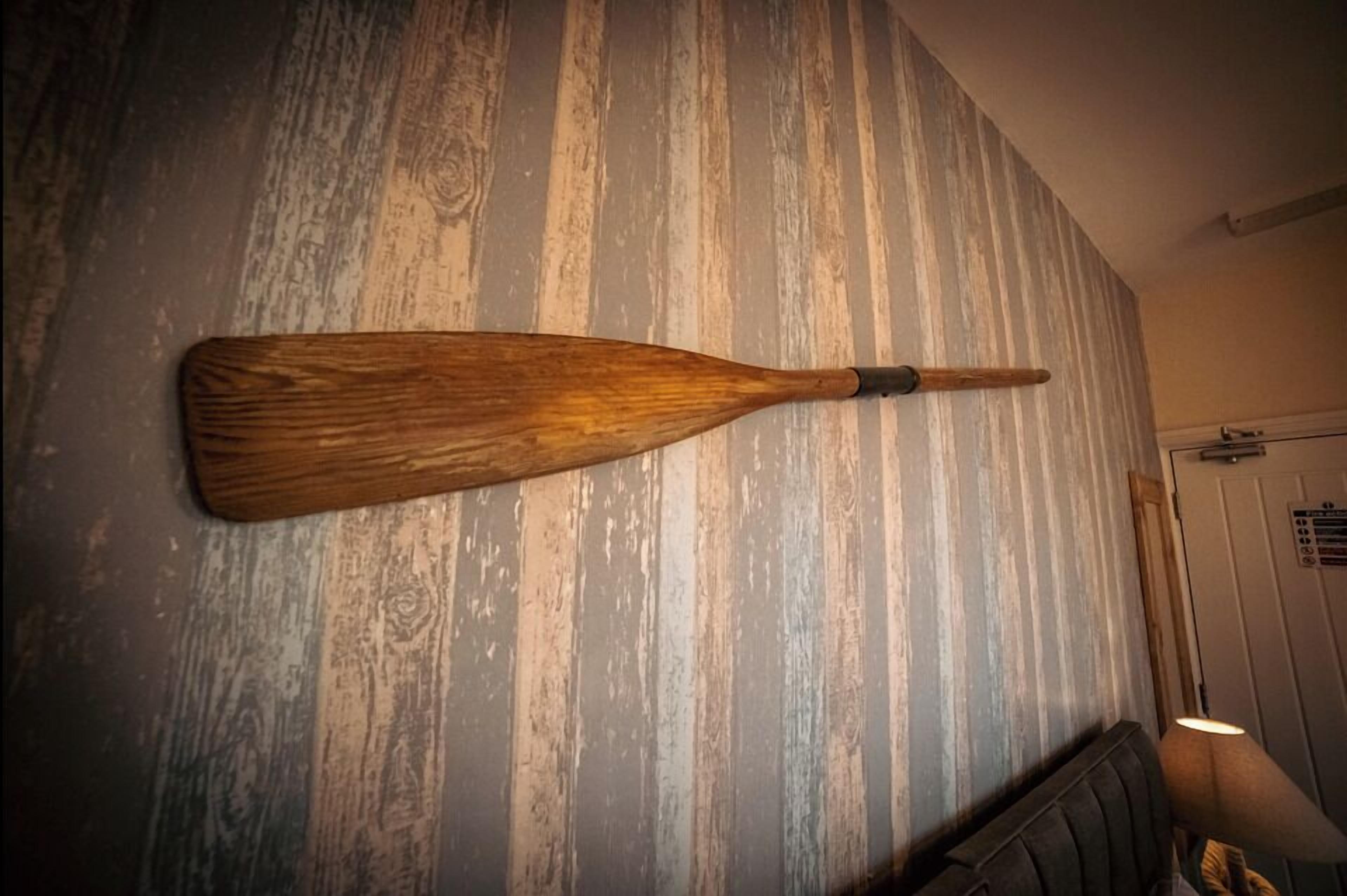 Une décoration murale représentant une pagaie en bois, dans une pièce dotée d’un mur en bois rustique et d’un canapé de couleur sombre.