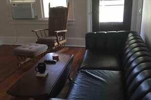 Un salón con un sofá de cuero negro, una silla de madera, un pequeño reposapiés y una mesa de centro con un control remoto y una taza.