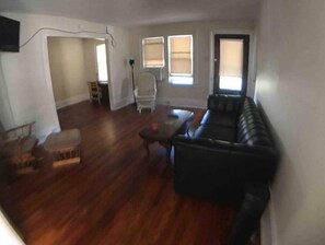 Una sala de estar con un sofá de cuero negro, una mesa de centro de madera, un sillón blanco y una pequeña mesa de madera. Hay dos ventanas con persianas, una puerta y una pared blanca.