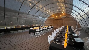 A long table set for a formal event inside a greenhouse with a wooden floor and white chairs.