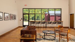 A spacious lobby with a wooden ceiling, a brown leather sofa, a round coffee table, and a large window offering a view of a red barn and trees.