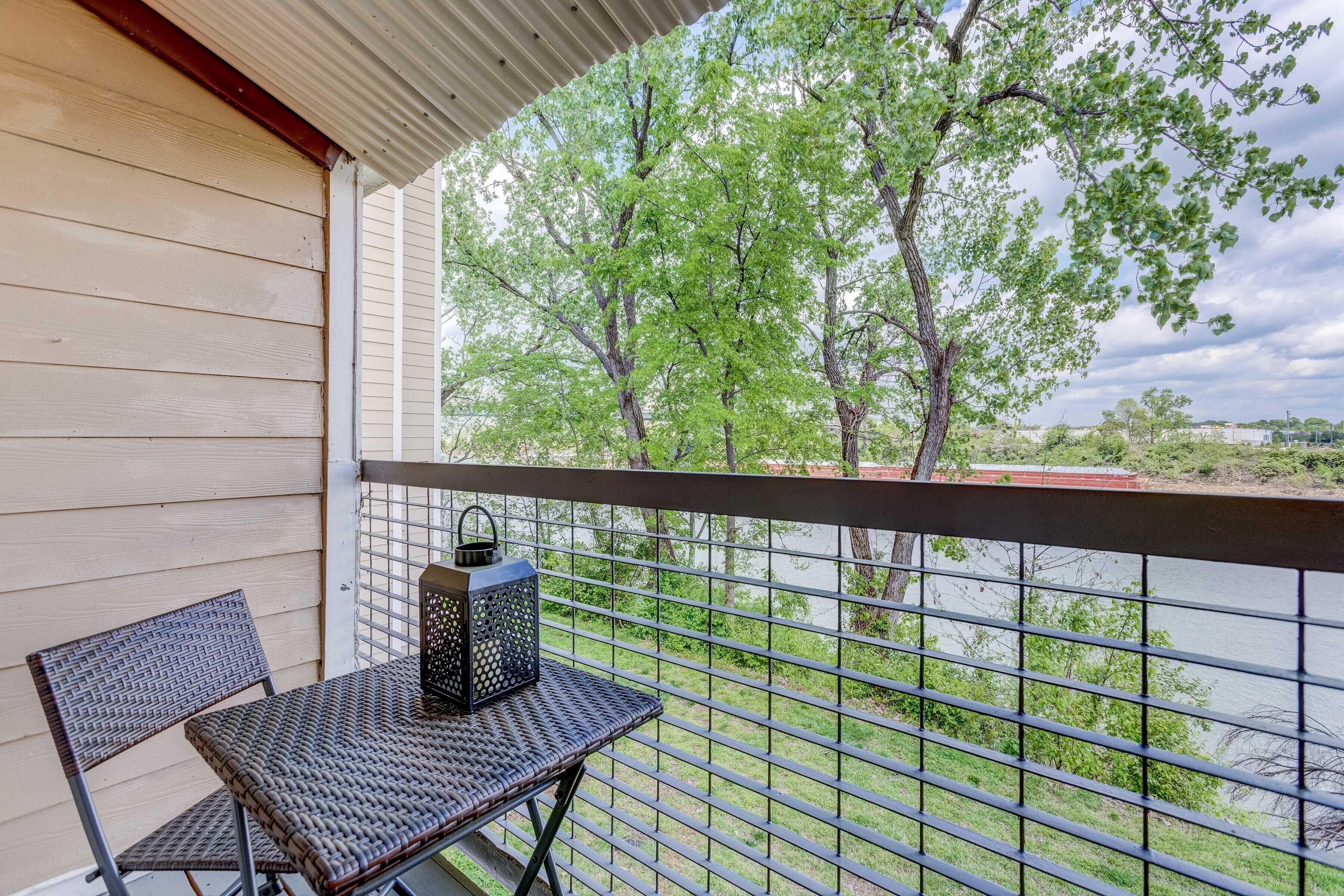 A balcony with a wicker table and chair, a lantern, and a view of a lake with trees.