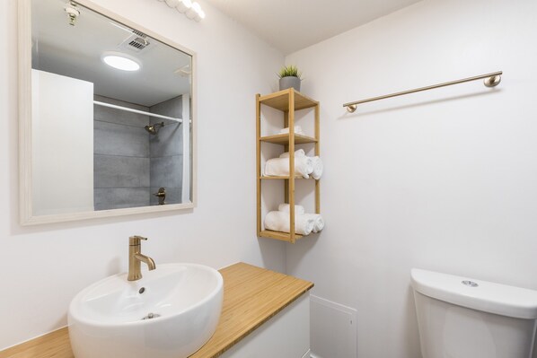 A bathroom with a white sink, a mirror, a wooden shelf with towels, and a toilet. - Modern Studio on I-Drive - pool + parking included (Orlando)