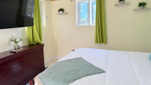 Ein Schlafzimmer mit Bett, Kommode, Fernseher, Klimaanlage, einem fenster mit Vorhängen und kleinen Topfpflanzen auf Regalen.