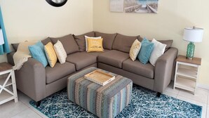 Ein Wohnzimmer mit einem braunen Ecksofa, einem gemusterten Hocker und einer Uhr an der Wand.