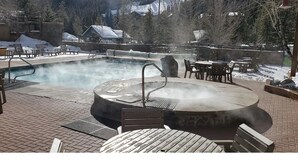 Un bain à remous entouré de meubles de jardin et d’une piscine, avec un sol et des arbres couverts de neige en arrière-plan.