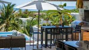 Um pátio externo com piscina, área de refeições e churrasqueira, com vista para o oceano.