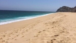 Uma praia arenosa com pegadas a levar à água, céu claro e um rochedo ao fundo.