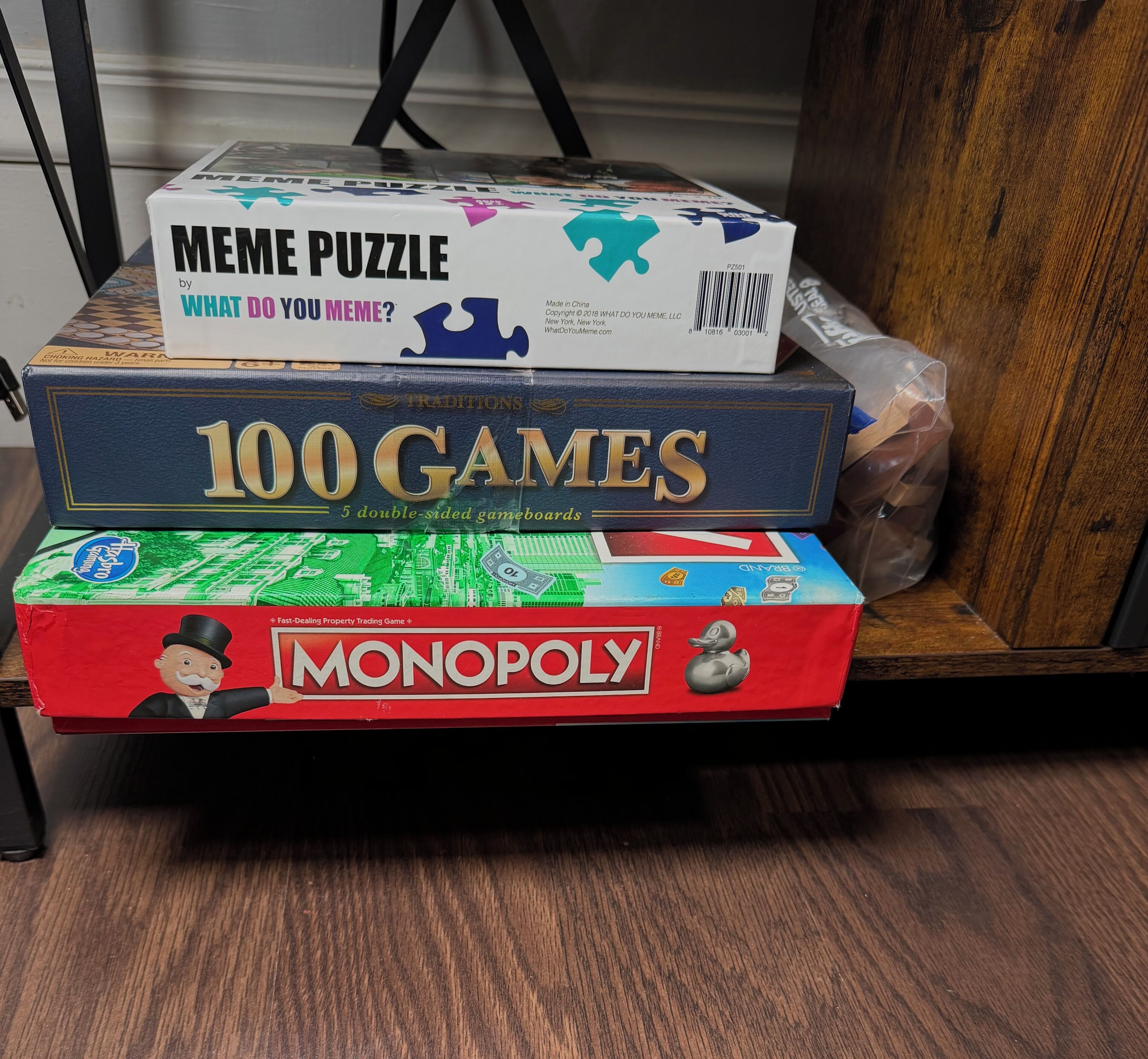 Three board games: 'Meme Puzzle,' '100 Games,' and 'Monopoly,' stacked on a shelf.