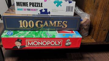 Three board games: 'Meme Puzzle,' '100 Games,' and 'Monopoly,' stacked on a shelf.