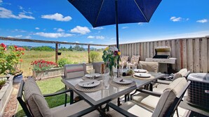 An outdoor patio with a table set for dining, surrounded by chairs, a grill, and a blue umbrella.