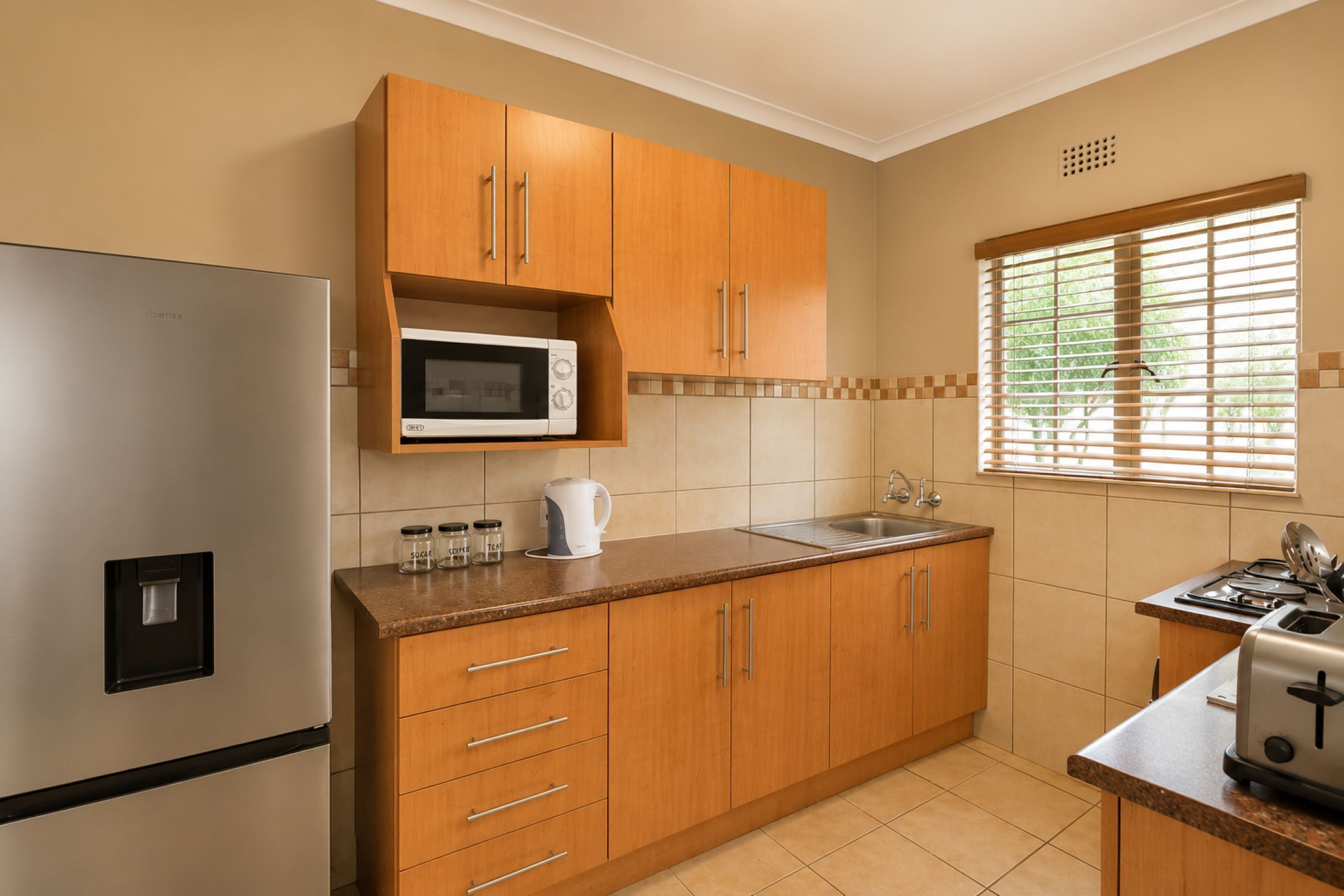 A kitchen with wooden cabinets, a stainless steel refrigerator, a microwave, a kettle, a sink, and a toaster.