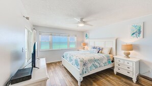 Un dormitorio con cama, mesitas de noche, ventilador de techo y ventana con vista al océano.