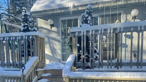 A hot tub with a wooden deck and railings, surrounded by snow-covered trees and a house with a balcony.