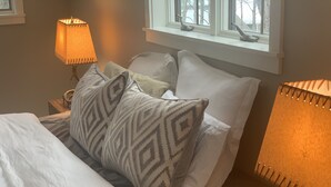 A neatly made bed with patterned pillows, a bedside lamp, and a window with a view of trees.