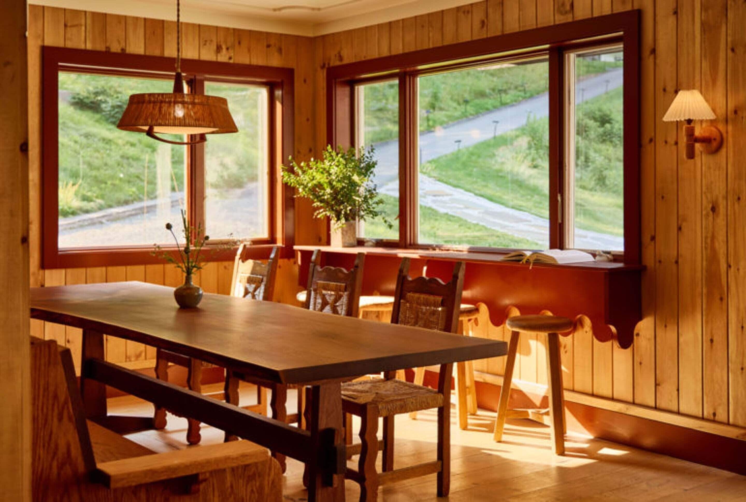 A wooden dining room with a large table, benches, and chairs. There are two windows with views of a road and greenery. A lamp hangs from the ceiling, and a plant is on the windowsill.