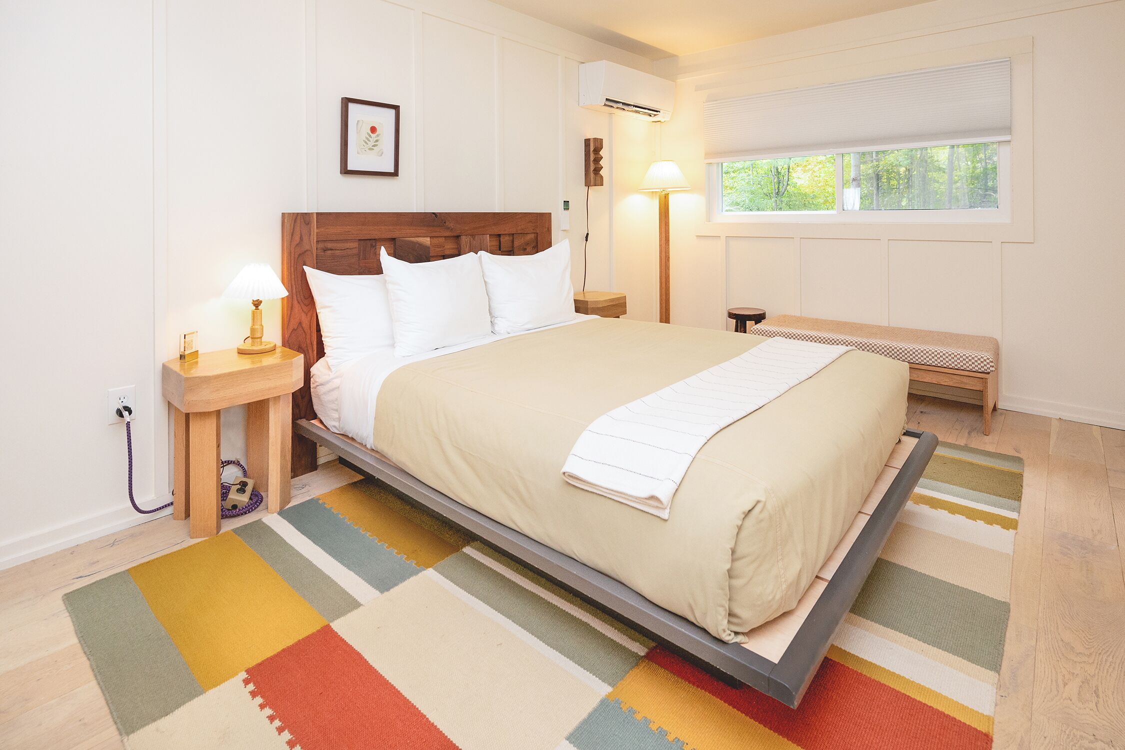 A neatly arranged bedroom with a bed, bedside tables, a nightstand, a bench, a window, and a wall-mounted air conditioner.