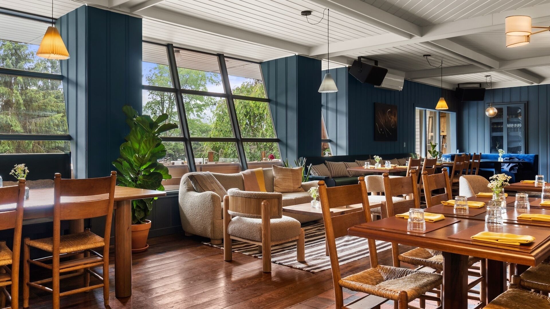 A spacious dining area with wooden tables and chairs, large windows offering a view of greenery, and a ceiling with exposed beams.