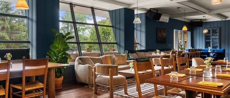 A spacious dining area with wooden tables and chairs, large windows offering a view of greenery, and a ceiling with exposed beams.
