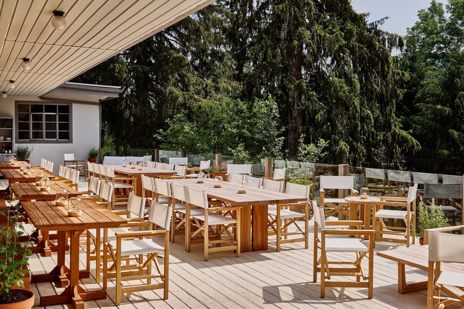Een buitenruimte met eettafels en stoelen van hout, omringd door groen en bomen.