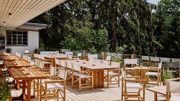 Een buitenruimte met eettafels en stoelen van hout, omringd door groen en bomen.